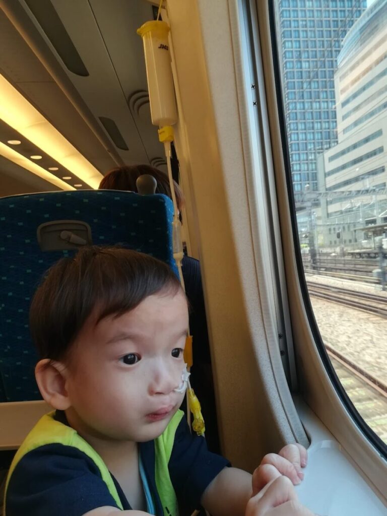 注入ボトルが新幹線のフックに吊り下げられている横で新幹線の窓から外を見る子ども