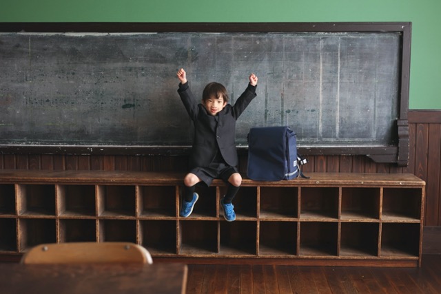 教室の後ろの棚の上に男の子が座っている。横にはランドセルがある