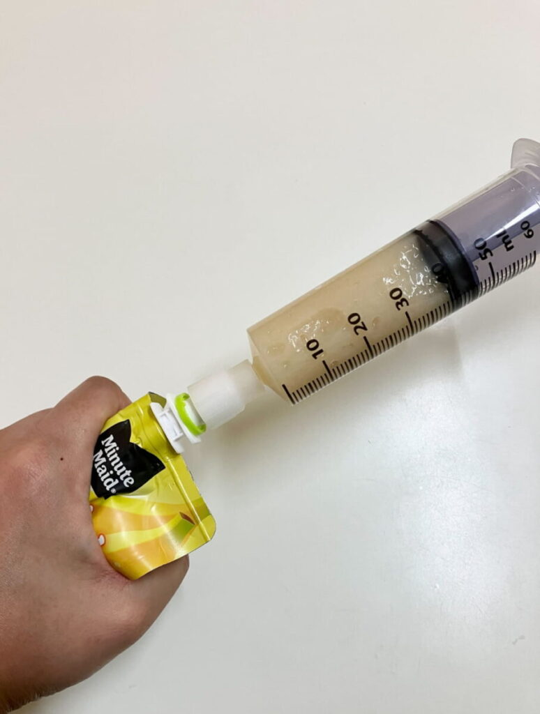 市販のパウチ型飲料と、注入用シリンジを接続するコネクタを接続して片手でパウチを握るだけでシリンジ内に中身を注入している様子