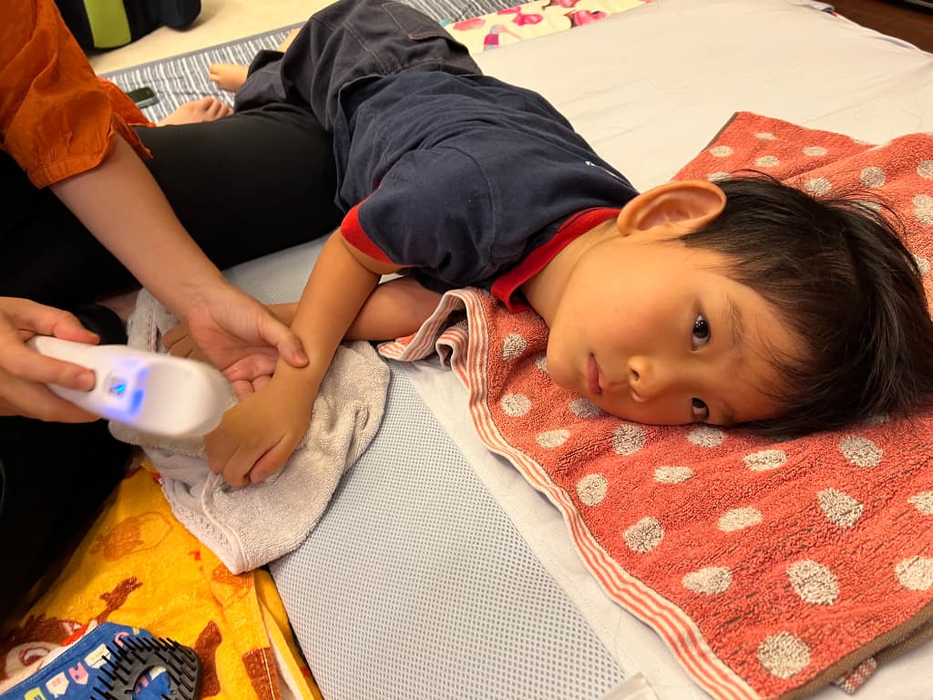 水玉模様のタオルの上に頭をのせて横になっている男の子。SUSUGUデバイスを手に当てて手を洗っている