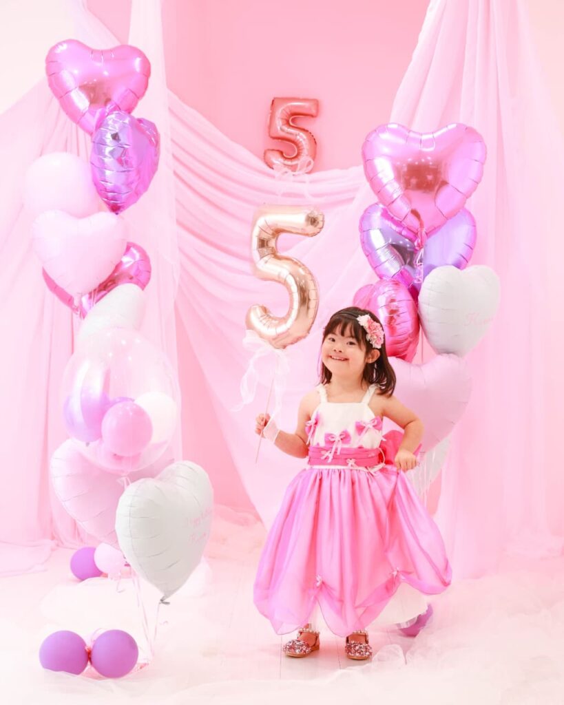 ドレスを着た女の子が、5の形をしたピンクの風船をもっている