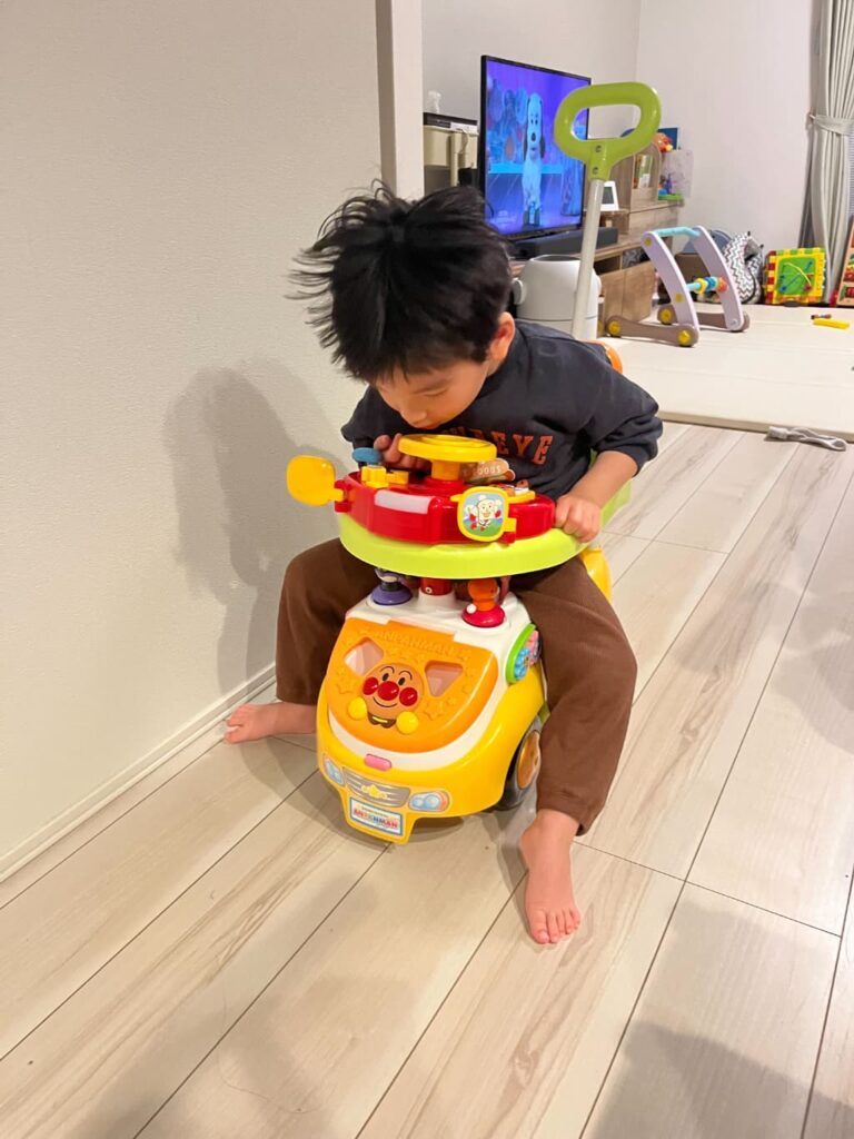 部屋の中で男の子が、アンパンマンのついた黄色いおもちゃの車に乗って遊んでいる