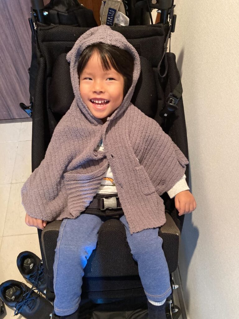 黒い子ども用車いすに座って笑っている男の子。茶色いニットのポンチョを着て、フードをかぶっている