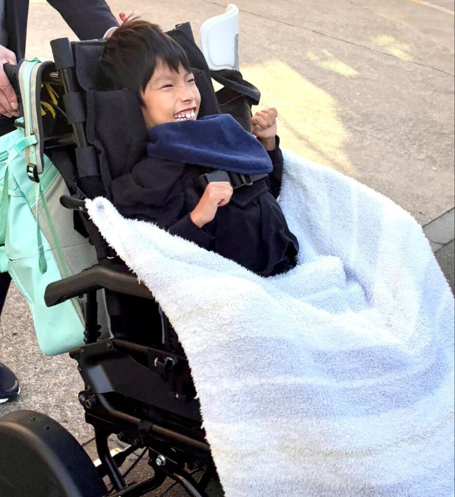 黒い車椅子に乗った笑顔の男の子が、外で車椅子を押してもらっているところ