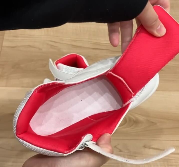 白い子ども用シューズの履き口を大きく開き、中のピンク色の中敷きと赤い内側部分が見える状態を手で持って撮影した写真