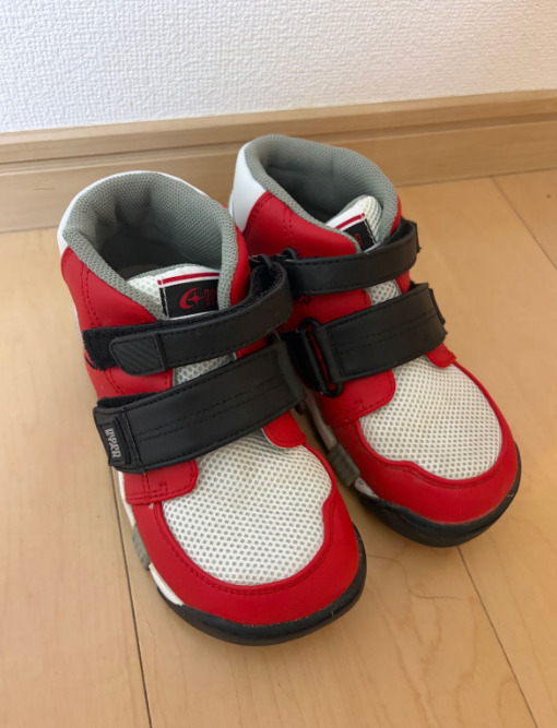 赤と白の配色の子ども用靴が、床の上に左右そろえて置かれている。面ファスナーのベルトが付いている