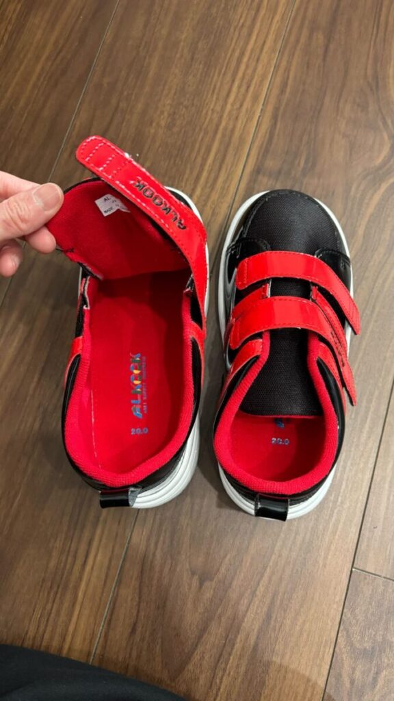 黒と赤の面ファスナー付き子ども用スニーカーを木の床の上で開いた状態で撮影した写真。履き口が大きく開き、着脱しやすい構造がわかる。