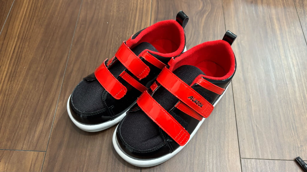 黒と赤の面ファスナー付き子ども用スニーカーを木の床に並べて撮影した写真。太いダブルベルトで足を固定できるデザインの靴。