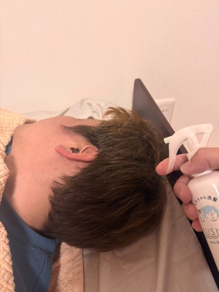 ベッドに横になっている人の頭部に、ミストタイプのシャンプーを吹きかけている様子。