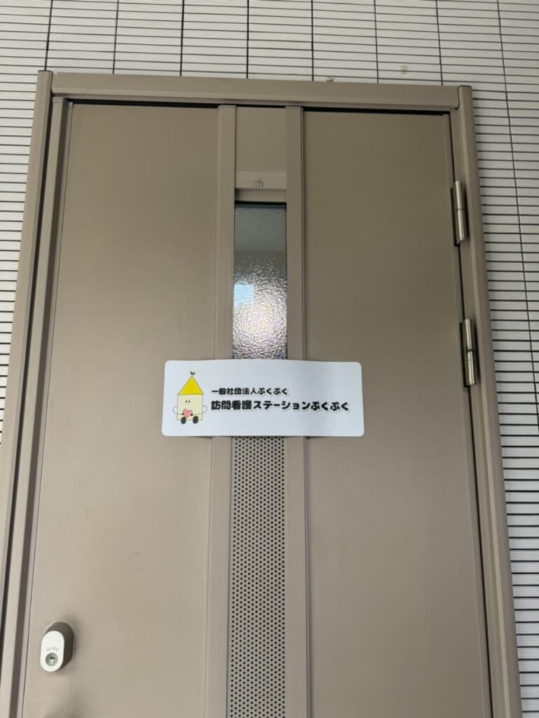 玄関ドアに訪問看護ステーションぷくぷくの施設名の看板が掲示されている様子。