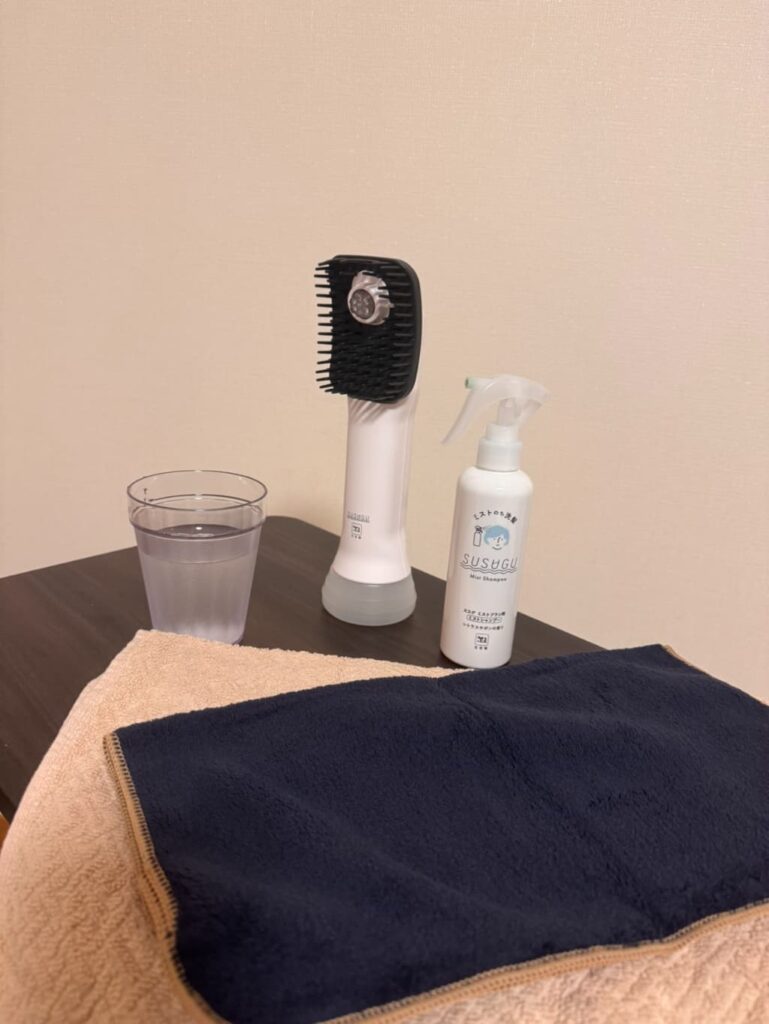 SUSUGU本体、ミストシャンプー、水の入ったコップとタオルがテーブルに並べられている様子。