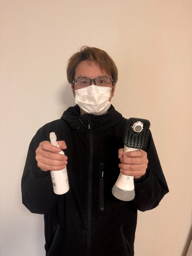 マスクを着用したスタッフが、ベッド上でも頭を洗えるブラシ付きの電動洗浄機（SUSUGU）とミストシャンプーを手に持って紹介している様子