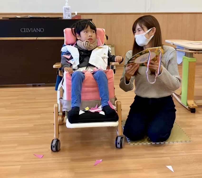 教室でピンク色の座位保持椅子に座った男の子とマスク姿の母親が隣に並んでいる様子。