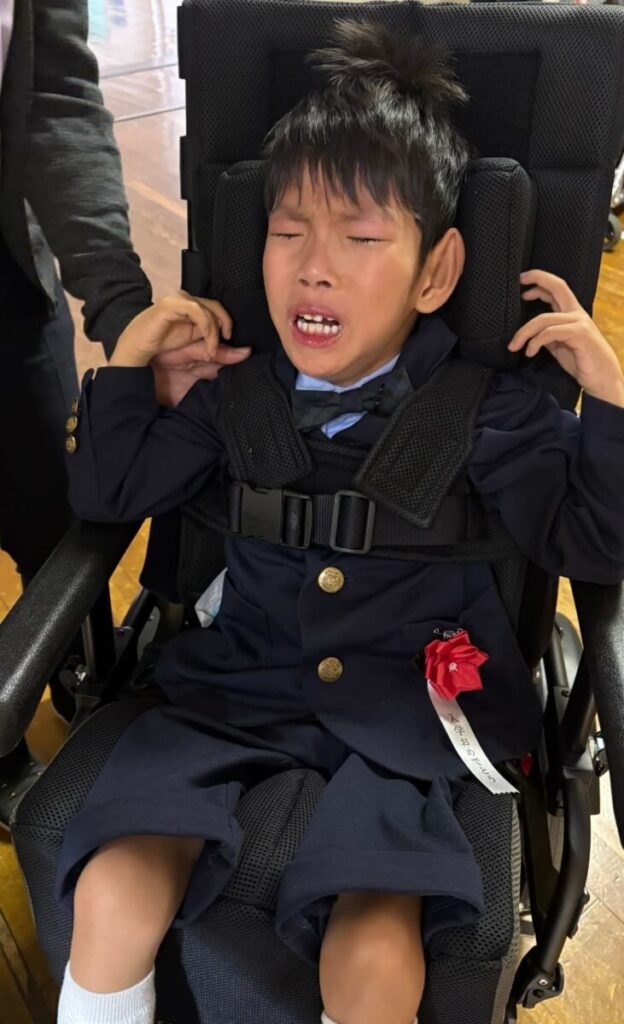 学校の屋内で、入学式用の制服を着た男の子が車いすに座り、式の時間を過ごしている様子。胸元には入学を示す赤いリボンが付いている