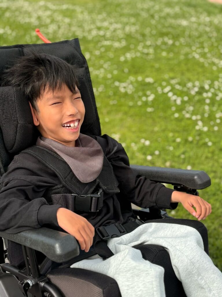 芝生と花が広がる屋外で、車いすに座った男の子が笑顔を見せながらリラックスして過ごしている様子