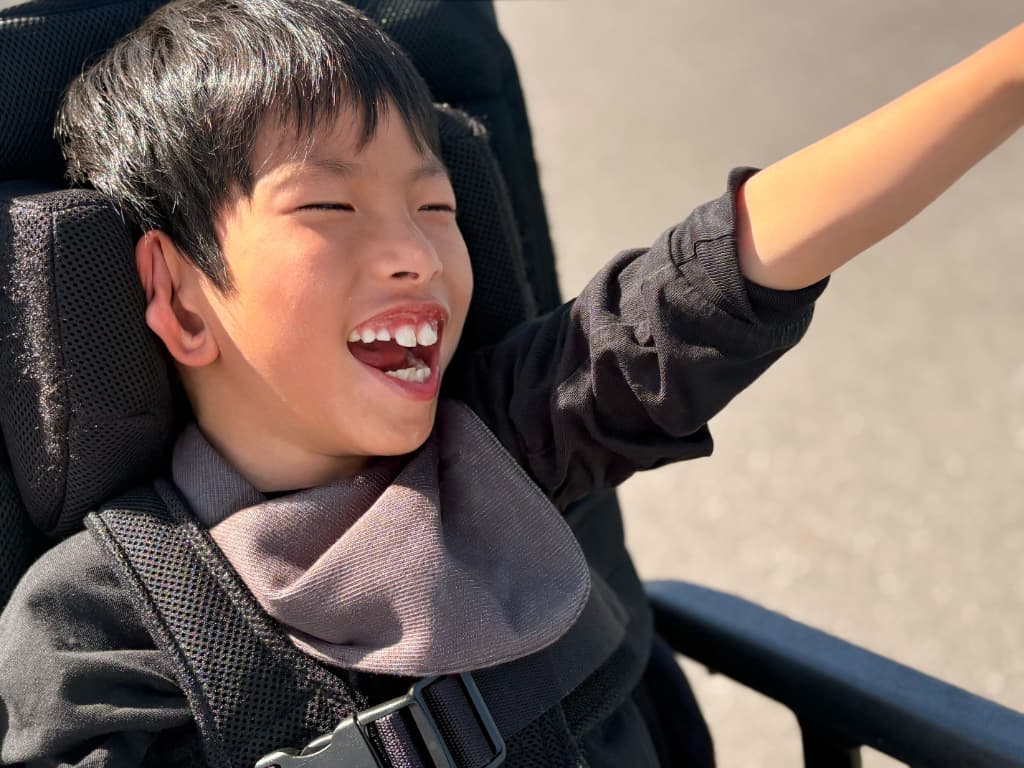 屋外で、車いすに座った男の子が手を前方へ伸ばしながら笑顔を見せ、外の時間を楽しんでいる様子