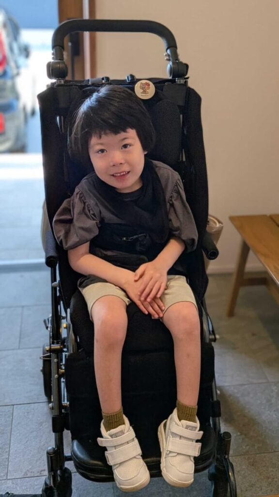 クリーム色のズボンとグレーのTシャツを着た笑顔の女の子が、玄関で黒いバギーに座っている。