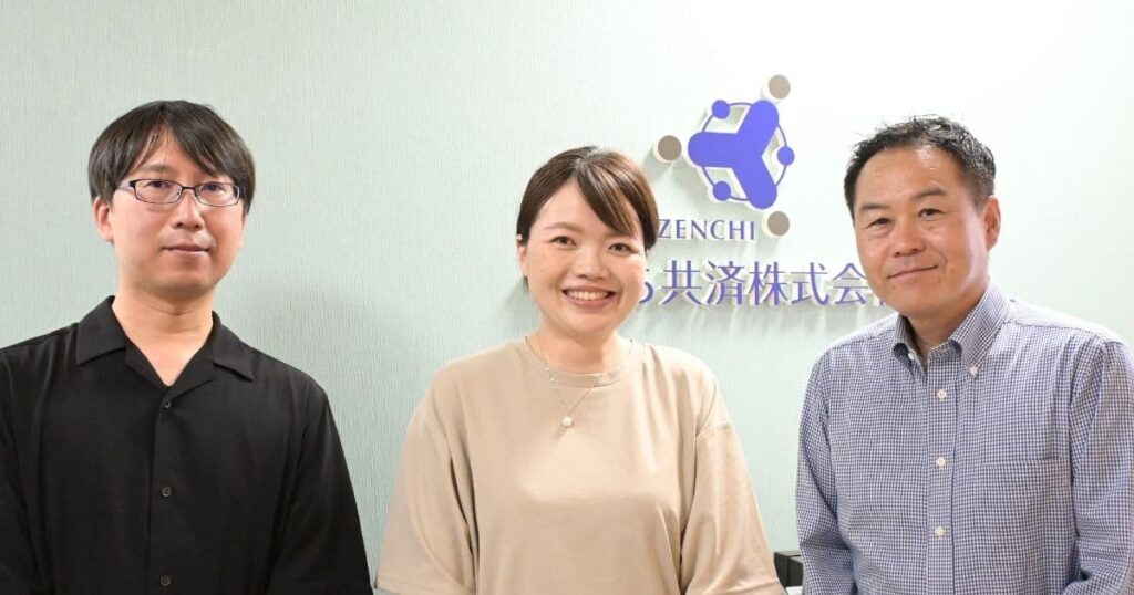 ぜんち共済株式会社の社員3名が会社ロゴの前でにこやかに立っている様子