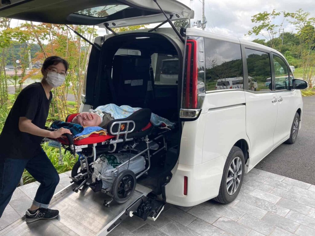 白い大型車両の後方からスロープを出して、障がい者用バギーに乗った子どもを車に載せている母親