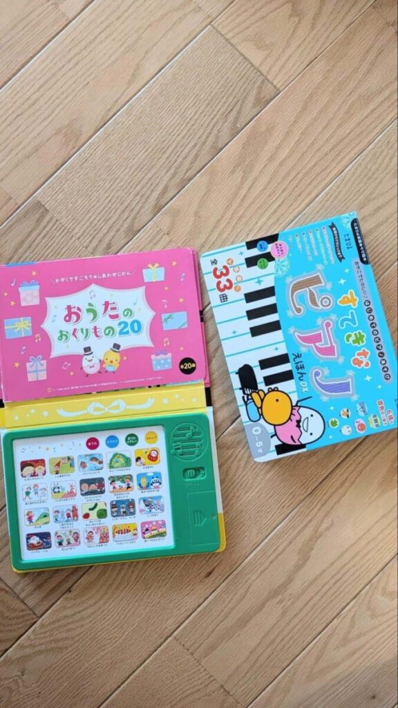 木目の床に並べた、ピンク色の「おうたのおくりもの20」と、鍵盤と音声ボタンが付いた水色の音楽絵本