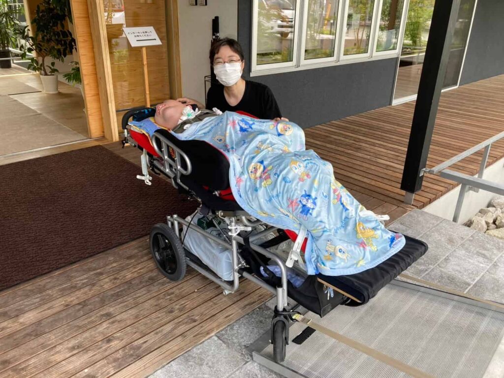 家の前で、障がい児用バギーに乗っている男性の後ろに、眼鏡をかけて微笑んでいる女性が寄り添っている様子