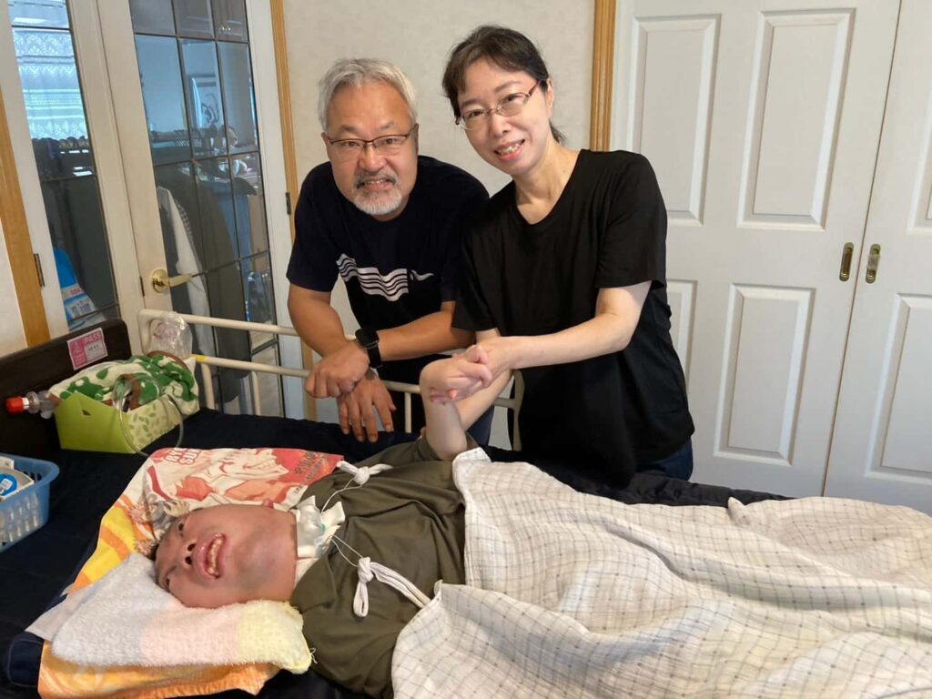 家の中で、電動ベッドの上に横たわる男性と、その両親の家族写真。母親は男性の手を握っており、母親の隣には父親が寄り添っている。