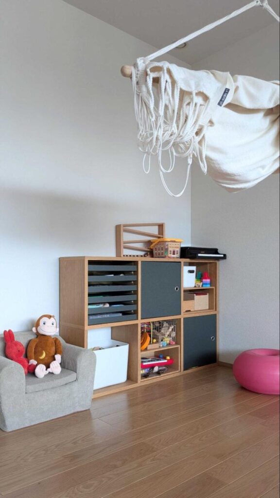 木製の収納棚の横に、サルと赤いゾウのぬいぐるみが座ったグレーの子ども用ソファ。天井には白い布製ハンモック、床にはピンクのバランスボールがある