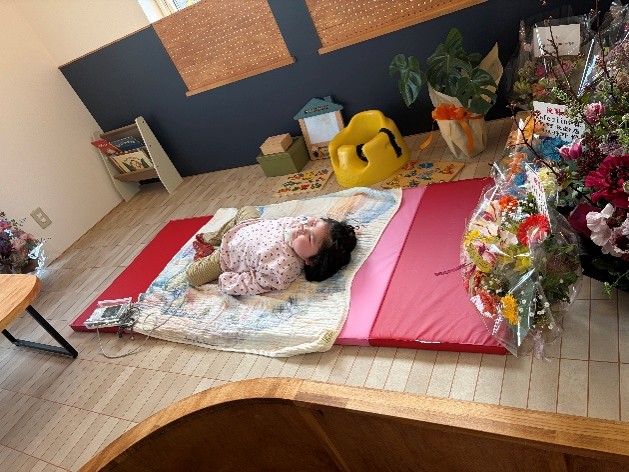 木目調の子上がりスペースに赤いマットが敷いてあり、女の子が横になっている。周囲にはお花やつみき、絵本などがある。