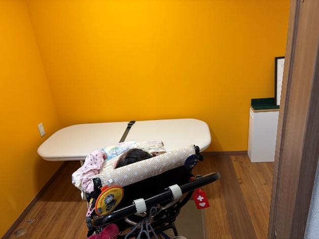 黄色い壁、茶色い木目の床の部屋にユニバーサルシートが置いてある。ユニバーサルベッドの前には、障がい児用バギーに乗った子どもがいる