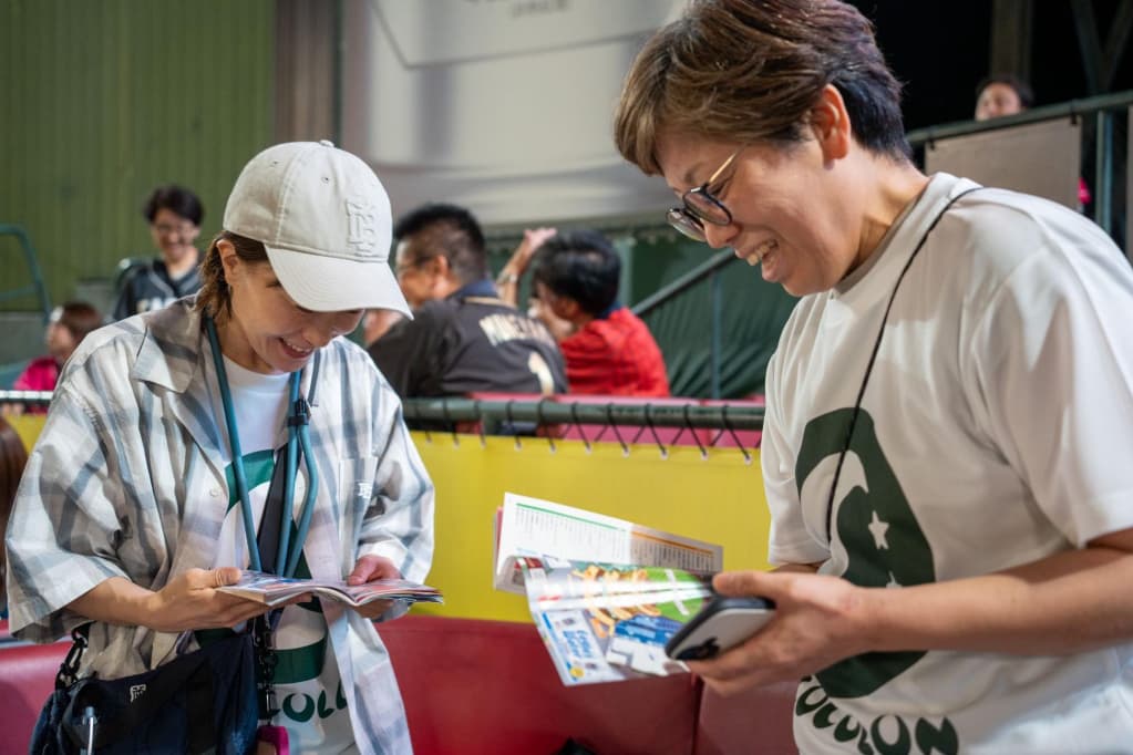 野球場で、白い帽子と青白のチェック柄シャツの女性、白いTシャツの男性が、イベントの冊子やチケットを手にして確認している様子。
