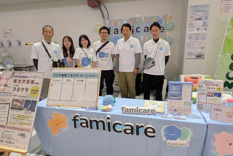 ファミケアの展示ブースで、スタッフ6人が並ぶ集合写真。前面にファミケアのロゴとキャラクターを配した水色のテーブルがあり、パウチ専用コネクタの紹介パネルや商品、サービス案内のチラシなどが並んでいる。