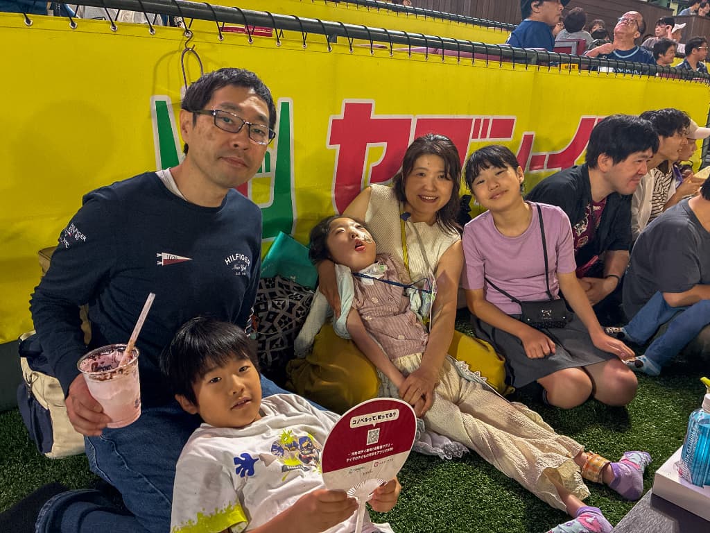 黄色いフェンス前の人工芝スペースで、黒い長袖の男性、ベージュの服の女性、ピンクの服の子どもたちが座る家族写真。