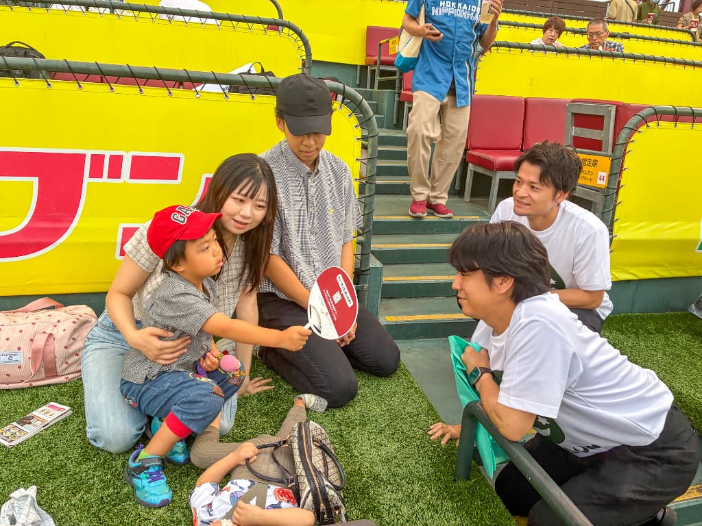黄色いフェンス前の人工芝で、赤いキャップの子どもを抱く女性と、白いTシャツの男性が座っている。手前では白いTシャツのスタッフがしゃがみ、子どもと目線を合わせている。