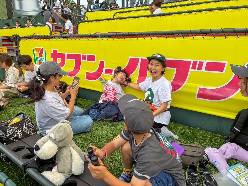 黄色いフェンス前の人工芝スペースで、白いTシャツと黒いキャップの女性が、赤白のボーダー柄の服を着た子どもを抱きながら笑っている。周囲には帽子をかぶった大人や子ども、バギー、ぬいぐるみがある。