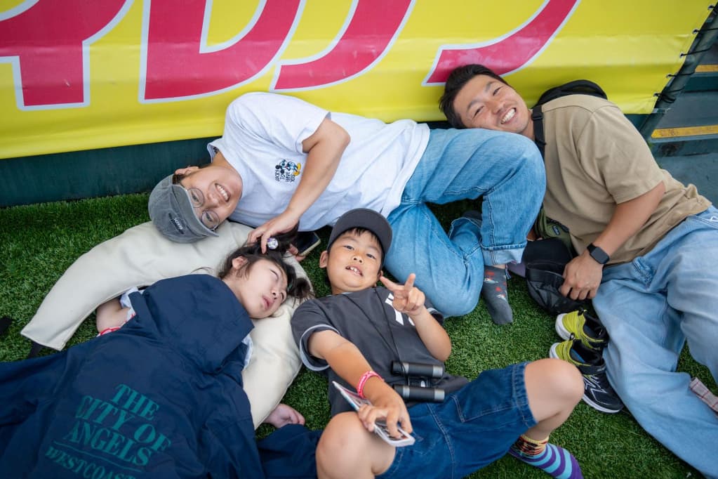 黄色いフェンス前の人工芝に、白いTシャツの男性、ベージュの服の女性、紺色の服の子ども、黒い服の子どもがクッションの上で横になっている様子。黒い服の子どもはカメラに向かってピースサインをしている。